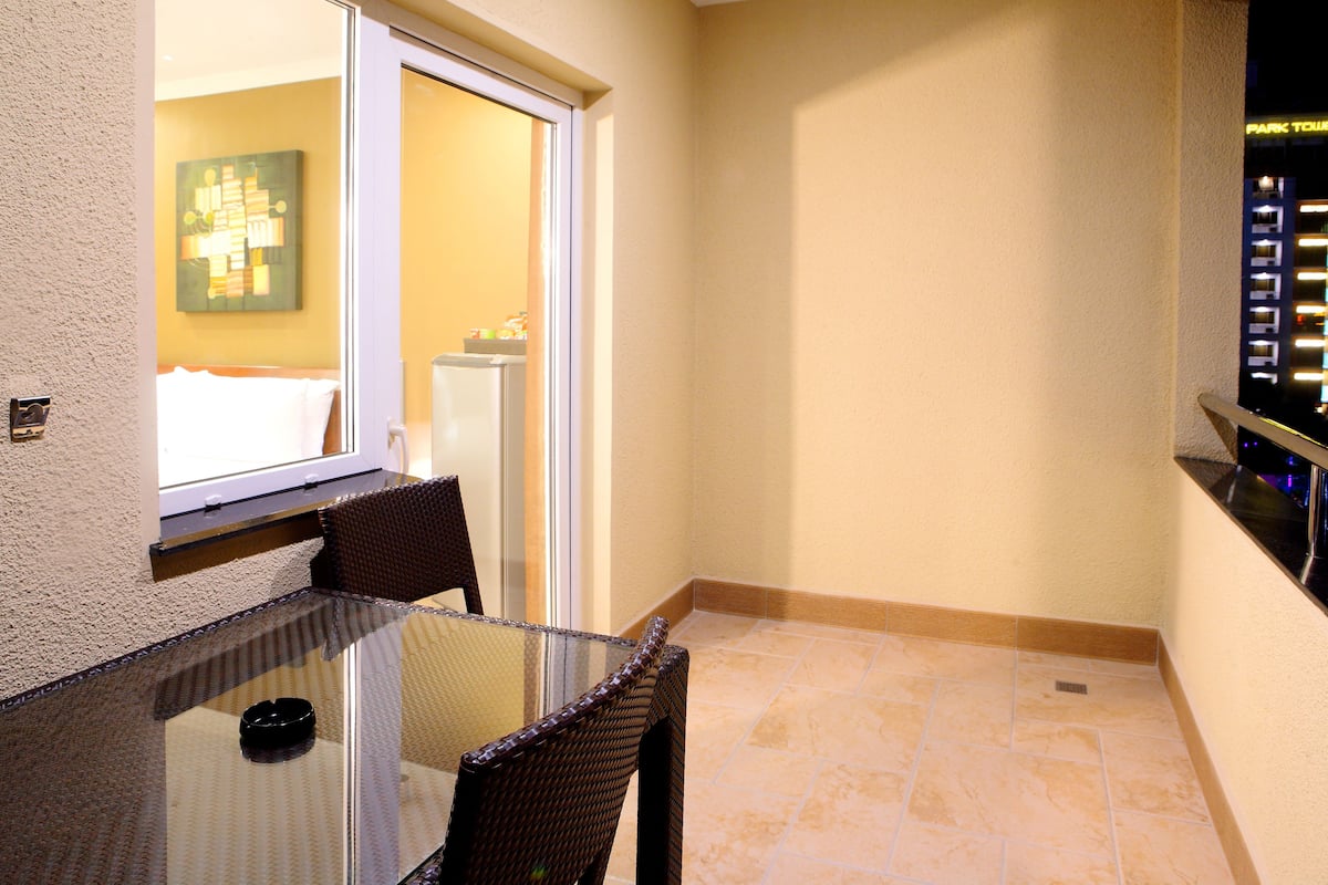A small balcony with a glass table and wicker chairs, a window with a view of a building, and a tiled floor.