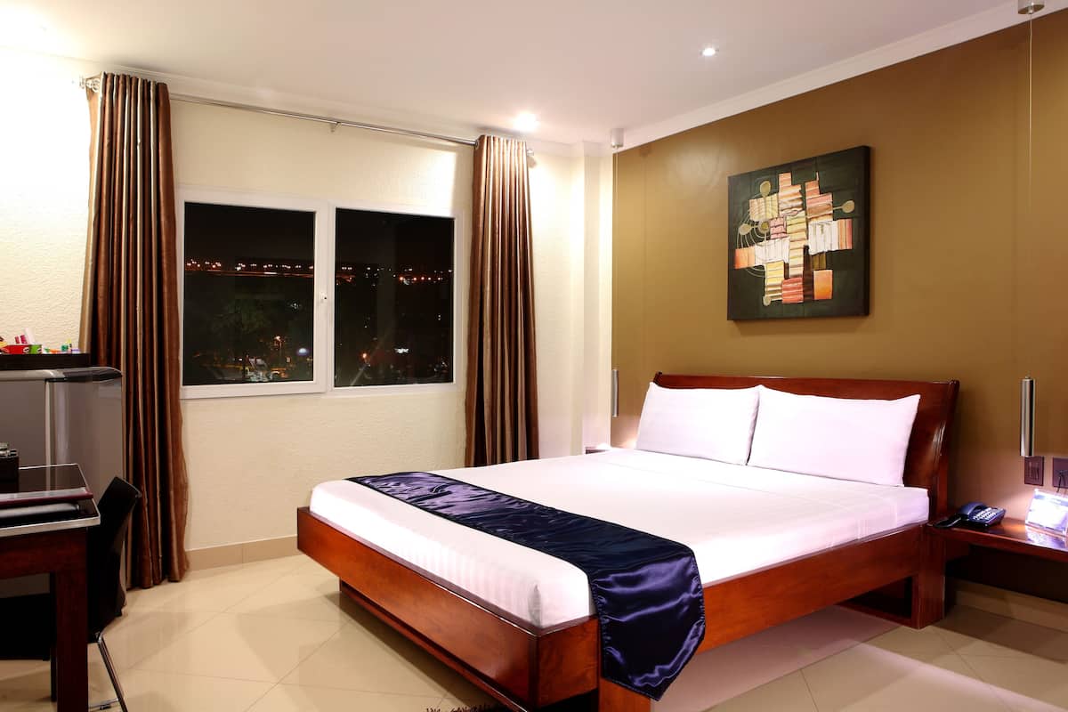 A hotel room with a large bed, a desk with a chair, a window with curtains, and a wall-mounted artwork.