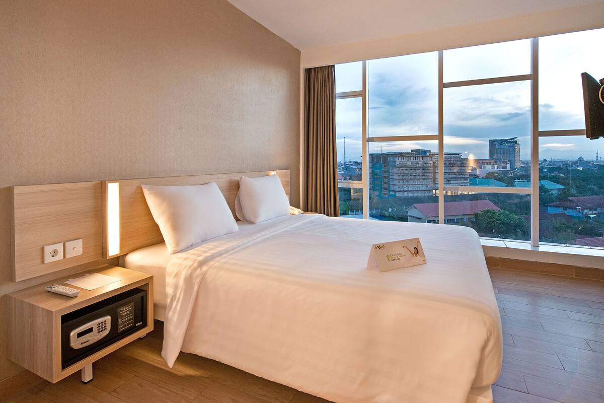 Quarto de hotel com uma cama grande, mesa de cabeceira com abajur e vista da cidade por grandes janelas.
