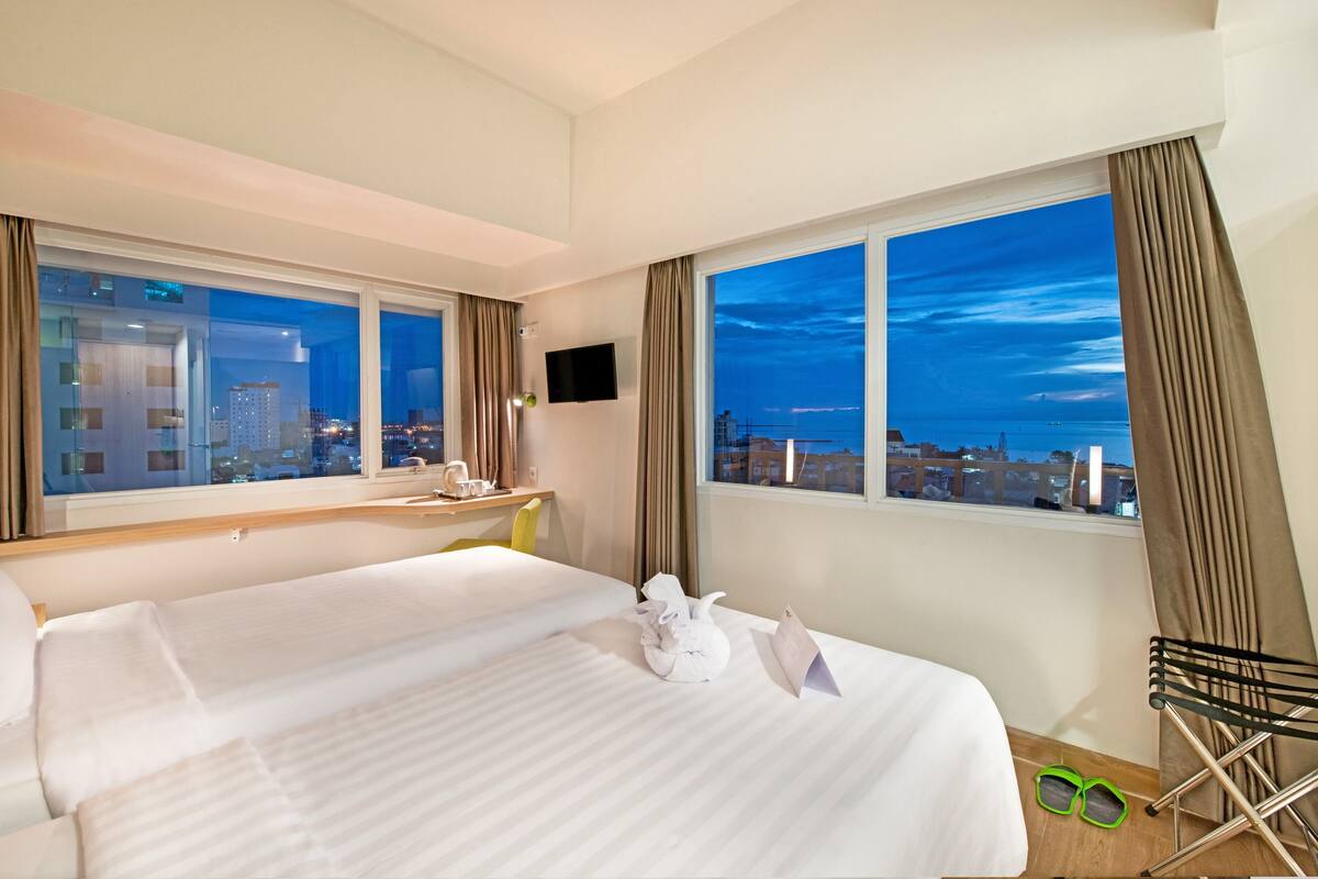 Quarto de hotel com uma cama grande, uma escrivaninha, uma cadeira e vista para a cidade à noite.