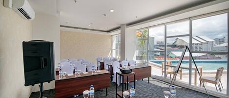 A conference room with a large table, chairs, and a projector. It has large windows with a view of a pool and buildings.