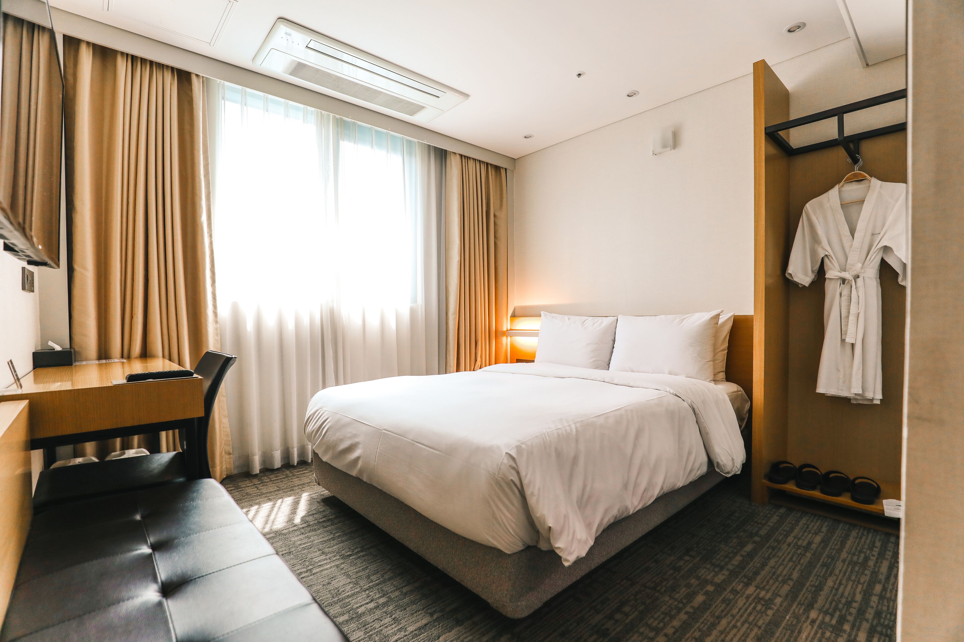 Una habitación de hotel con cama, escritorio, banco, bata colgada en la pared y zapatero.