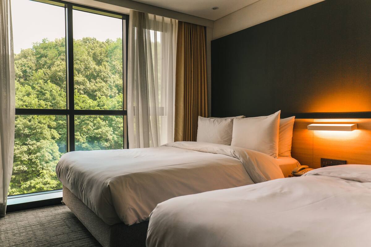 Une chambre d’hôtel avec deux lits, une grande fenêtre avec des rideaux, une lampe de chevet et un téléphone sur la table de chevet.