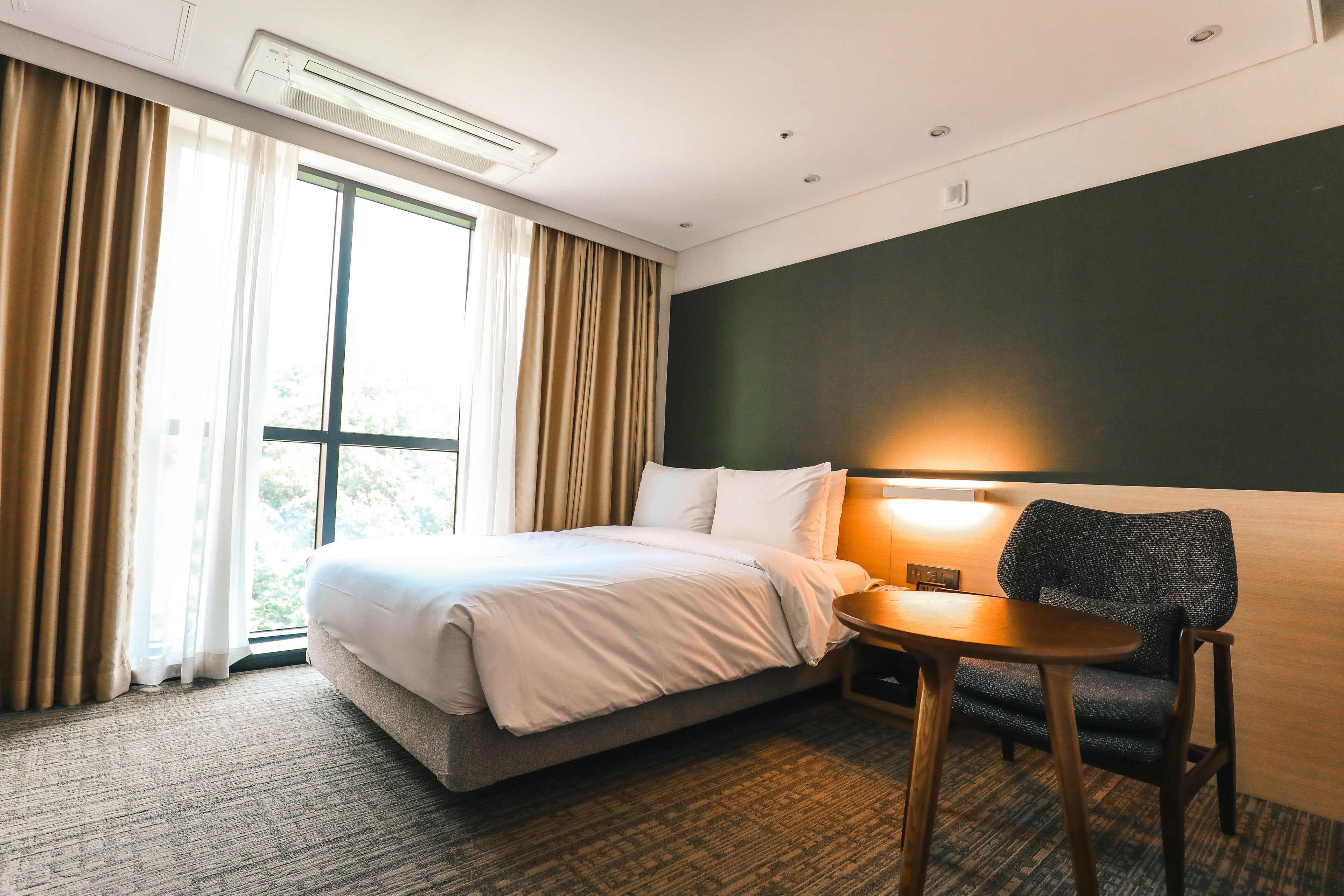 A hotel room with a bed, a small table, a chair, and a window with curtains.