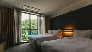 A hotel room with two beds, a large window with curtains, a wall-mounted lamp, and a view of greenery outside.