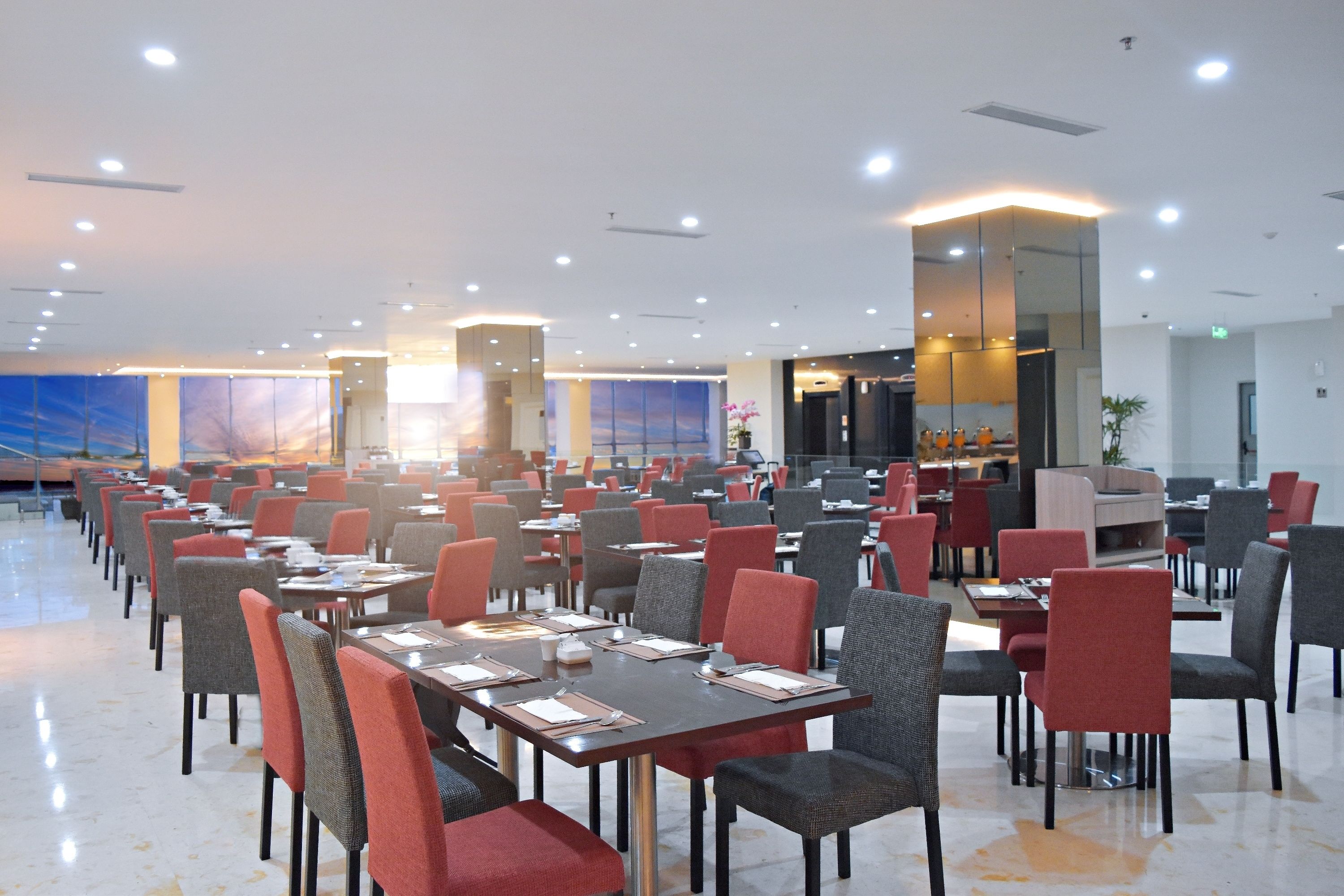 A spacious dining area with multiple tables and chairs, well-lit with ceiling lights, and a large window with a view of the sky.