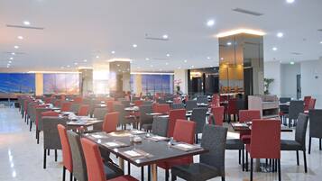 A spacious dining area with multiple tables and chairs, well-lit with ceiling lights, and a large window with a view of the sky.