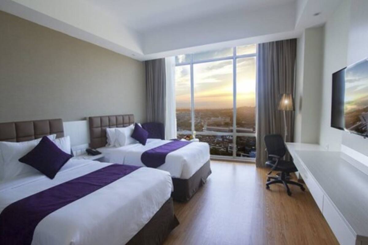 Habitación de hotel con dos camas, un ventanal grande con vistas a la ciudad, un escritorio y una silla.