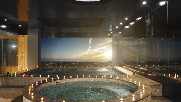 Een wellnessruimte met een grote ronde hottub, omringd door kaarsen, een houten sauna en een groot scherm waarop een zonsondergang te zien is.