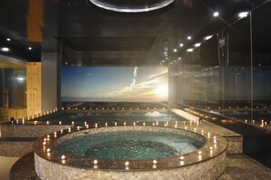 Een wellnessruimte met een grote ronde hottub, omringd door kaarsen, een houten sauna en een groot scherm waarop een zonsondergang te zien is.