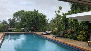 Una piscina rectangular con borde de azulejos, rodeada por una terraza de madera. Hay dos sillas reclinables y una pequeña mesa con una silla. La piscina está al lado de un edificio con techo blanco y está rodeada de abundante vegetación y árboles.