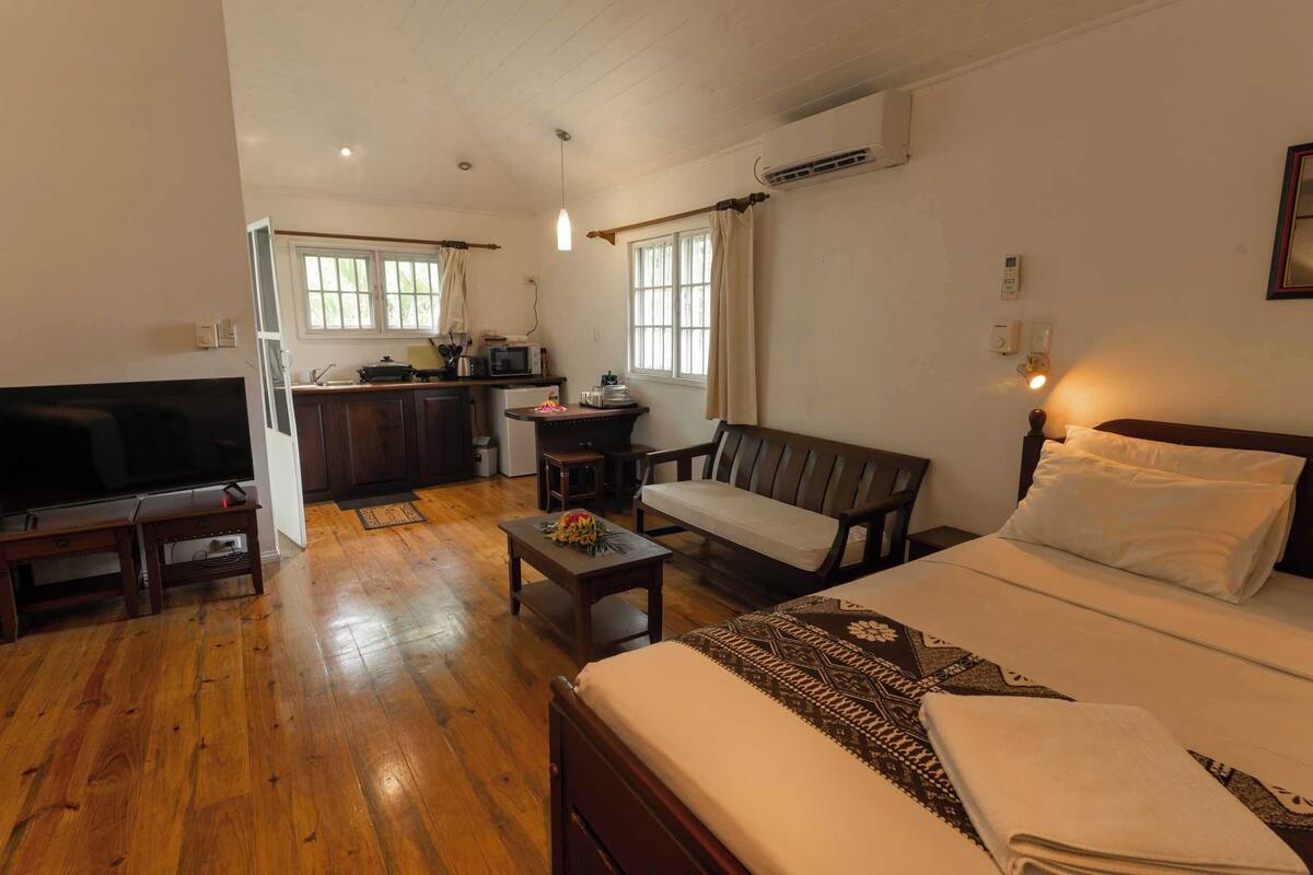 A compact hotel room with a bed, a sofa, a TV, a small kitchen area, and a dining table.
