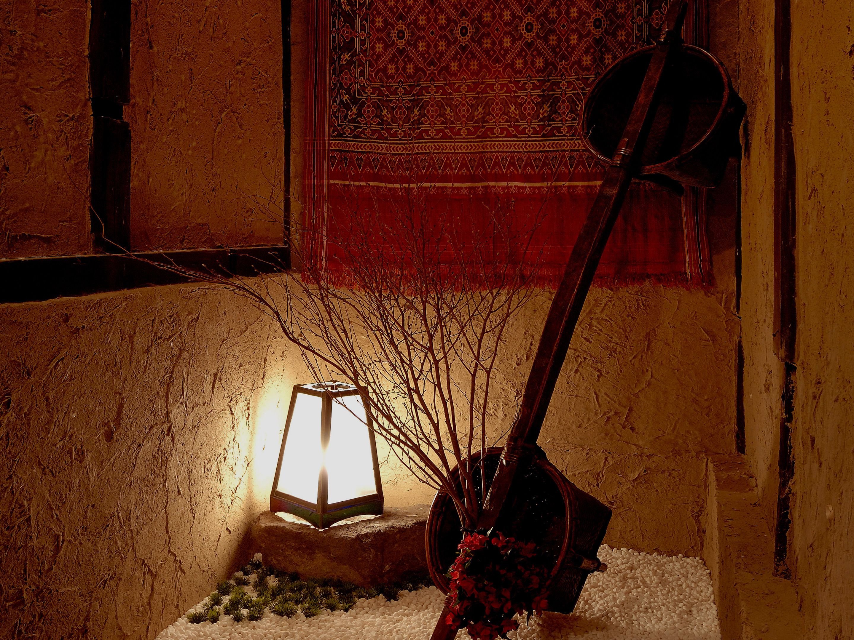 A lantern on a stone base with a flower arrangement, set against a textured wall with a patterned tapestry in the background.