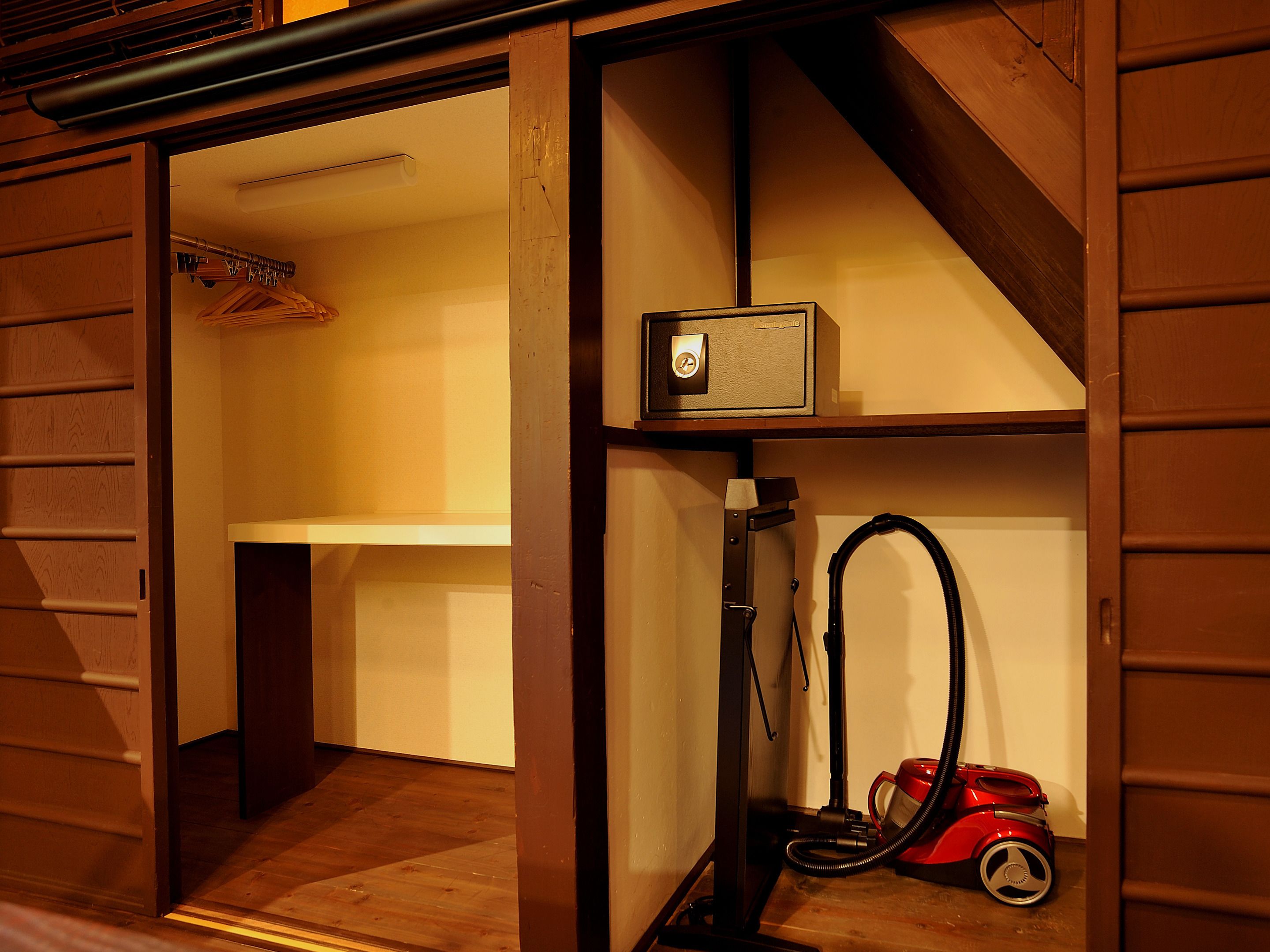 Habitación pequeña con piso de madera, un estante blanco y un aspirador rojo, además de una caja fuerte.