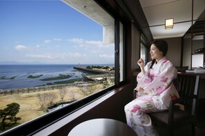 A room with a large window offering a view of a coastal area, a wooden chair, and a small table. - Hotel Nampuro (Shimabara)