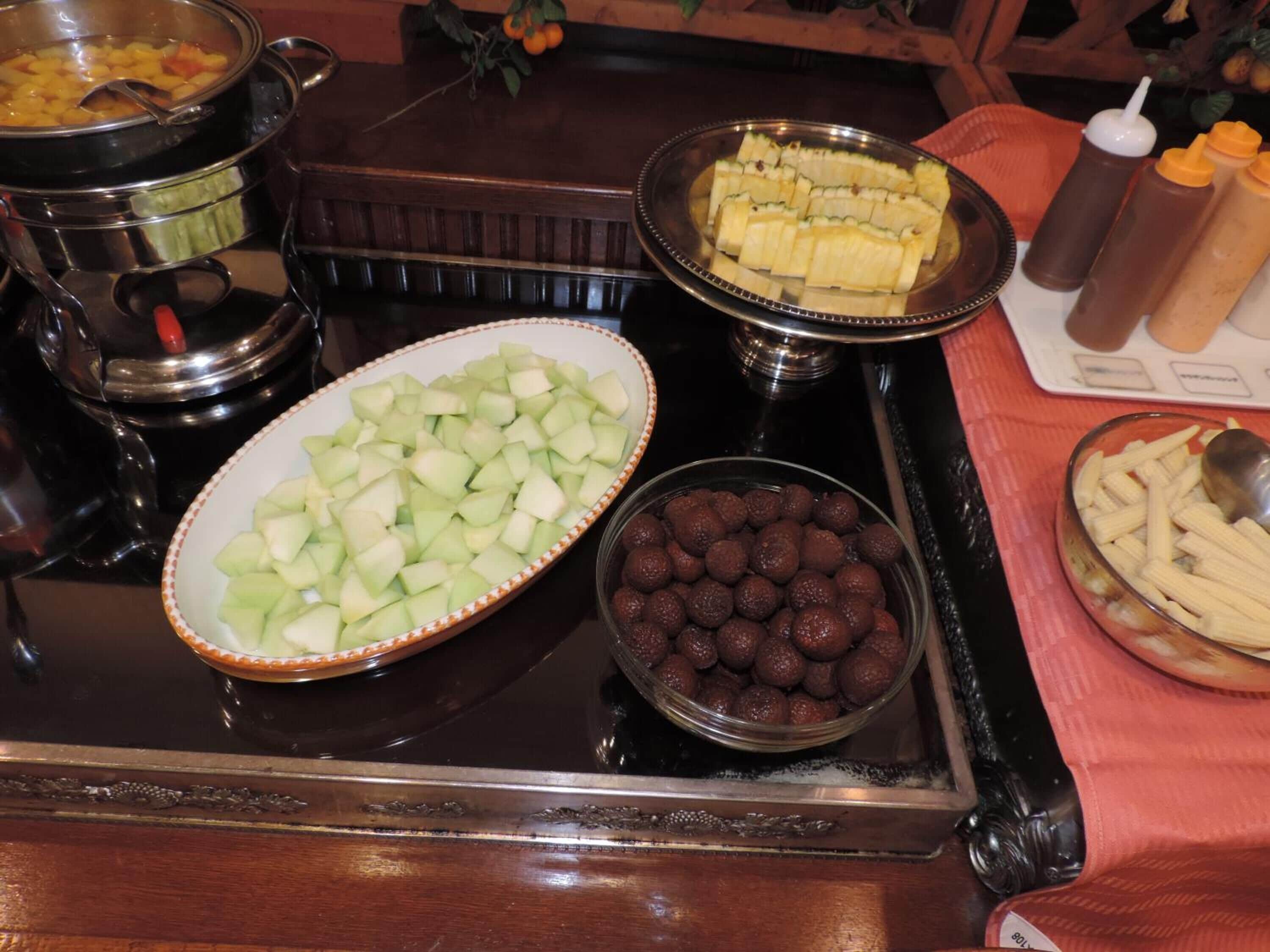 A buffet table with various food items including a large bowl of cubed fruit, a dish of sliced fruit, and a bowl of chocolate balls.