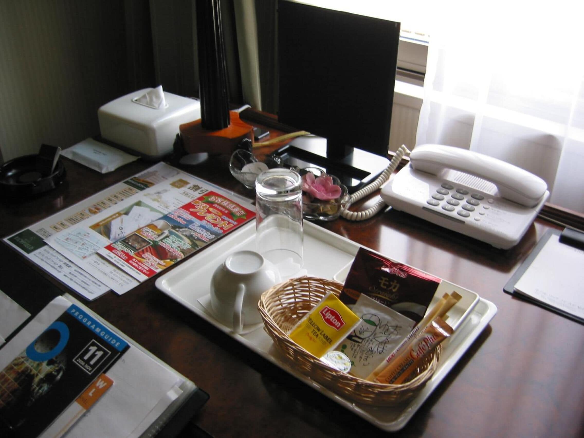 Een hotel bureau met een menukaart, een glas, een mandje met Lipton thee en koffie, een tissuedoos, een telefoon en een programmaboekje.