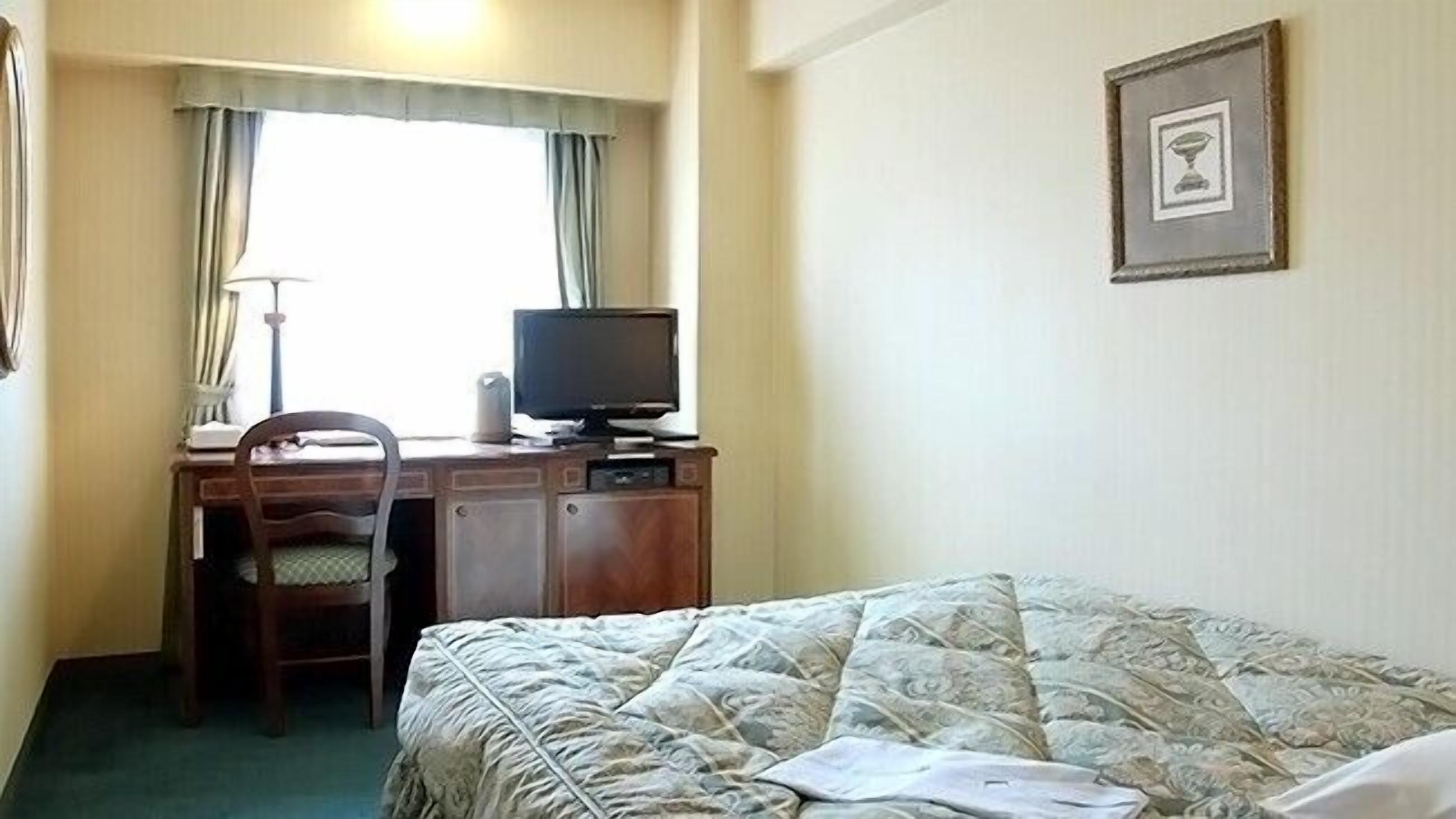 A hotel room with a bed, a desk with a computer, a chair, a window with curtains, and a framed picture on the wall.