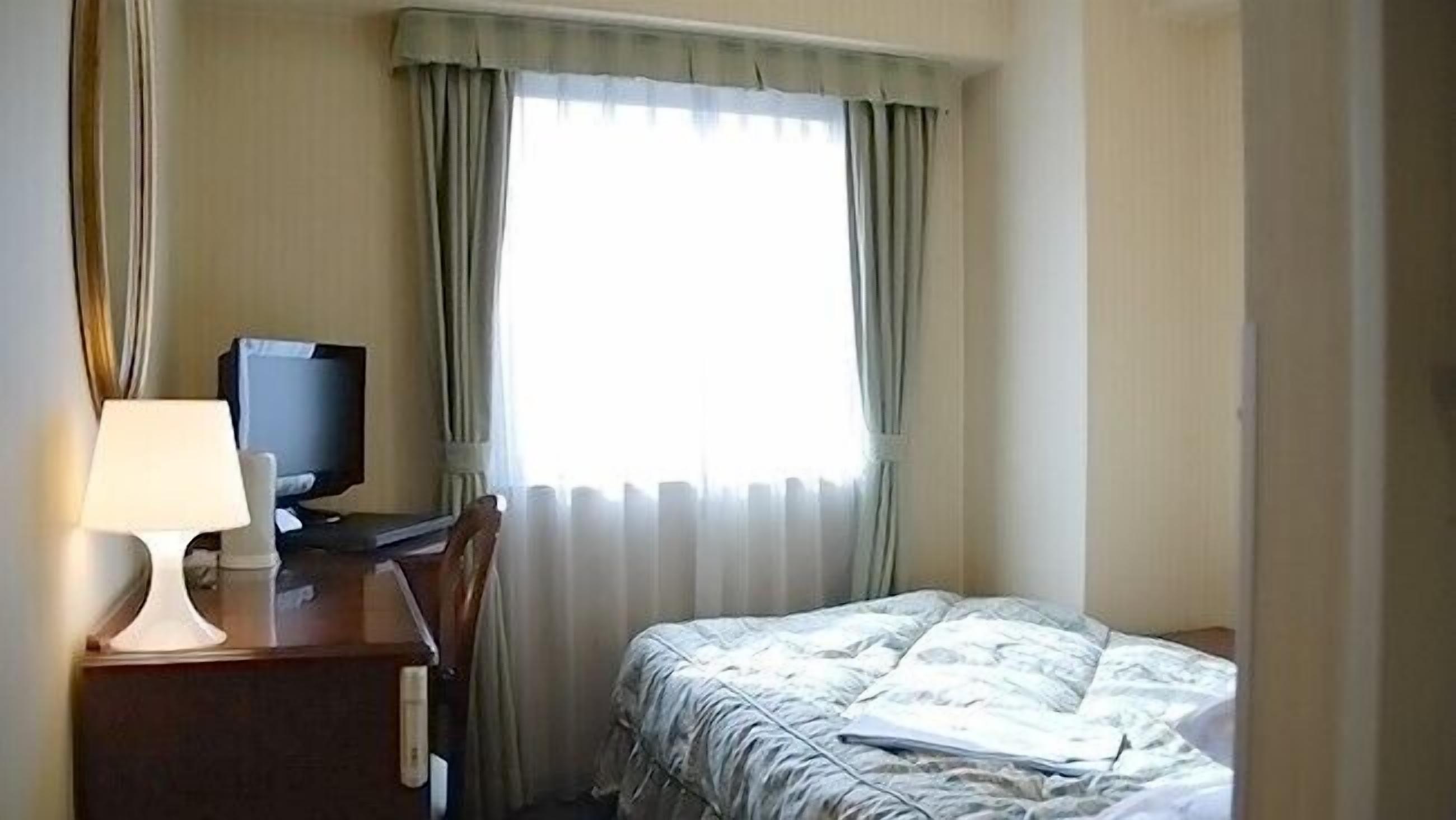 A hotel room with a bed, a desk with a television, and a window with curtains.