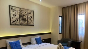 Quarto de hotel com uma cama grande, duas almofadas, cabeceira, mesa de cabeceira, uma cadeira, janela com cortinas e uma obra de arte na parede.