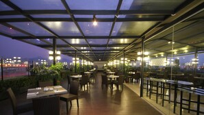 Um espaço de refeições interno com mesas e cadeiras, um guarda-corpo de vidro e vista para a cidade ao entardecer.