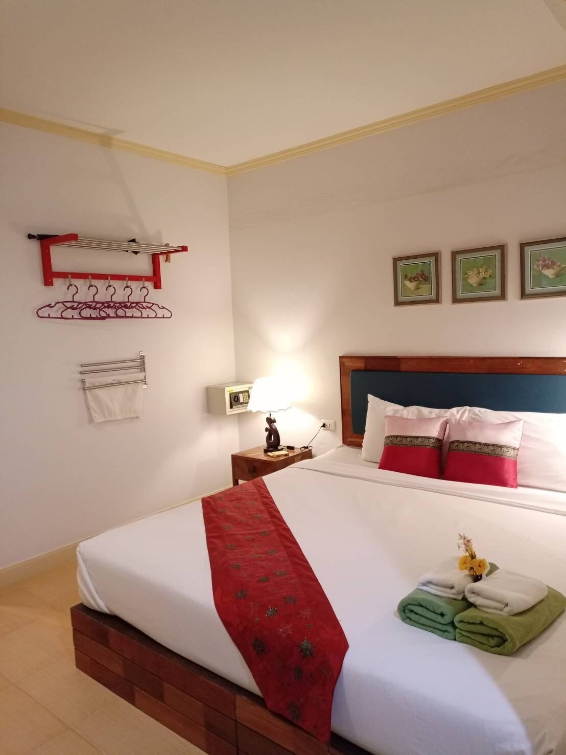 A neatly made bed with a red decorative cloth, a bedside lamp, and a wall-mounted drying rack with clothes.