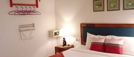 Una cama bien hecha con un cubrecolor rojo, una lámpara de noche y un tendedero de pared con ropa.