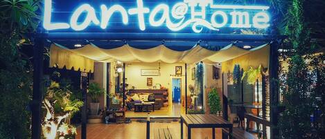A cozy, well-lit home with a neon sign reading "LantaHome." It features a welcoming entrance with a covered patio, potted plants, and a comfortable seating area.
