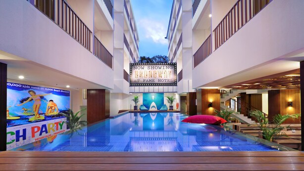 Un vestíbulo de hotel con una piscina central, un cartel que dice 'Broadway en Fame Hotel' y un cartel de evento llamado 'Beach Blanket'.