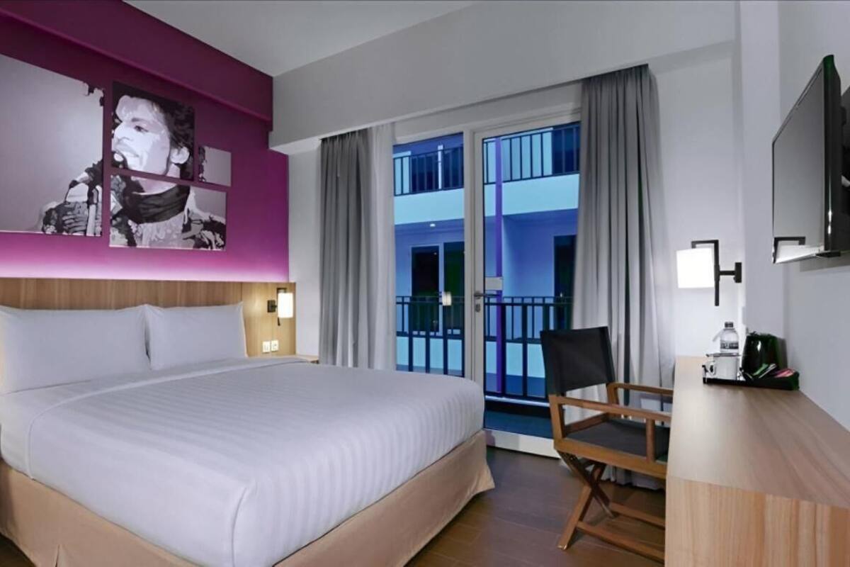 A hotel room with a large bed, a desk with a chair, a TV, and a balcony with curtains.