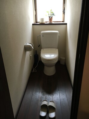 A small, narrow bathroom with a toilet, a window, and a pair of slippers on the floor. - Ryoso Kawaguchi (Hatsukaichi)
