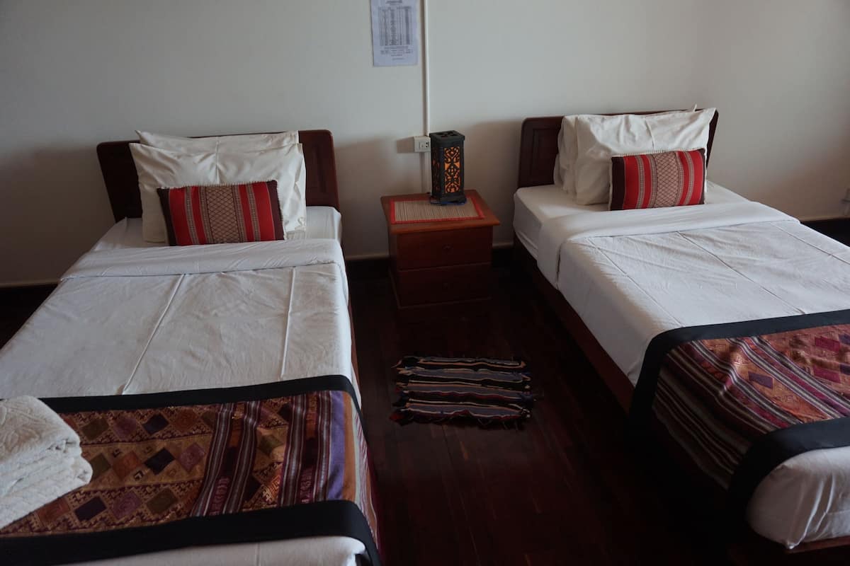 Two single beds with white linens and red and brown patterned pillows, a wooden nightstand with a decorative lantern, and a patterned rug on the wooden floor.
