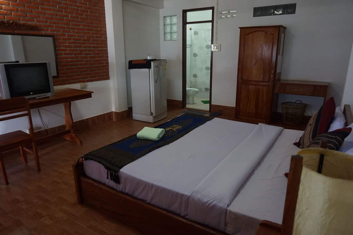 A bedroom with a bed, a desk with a television, a refrigerator, a wardrobe, and a bathroom visible through an open door.