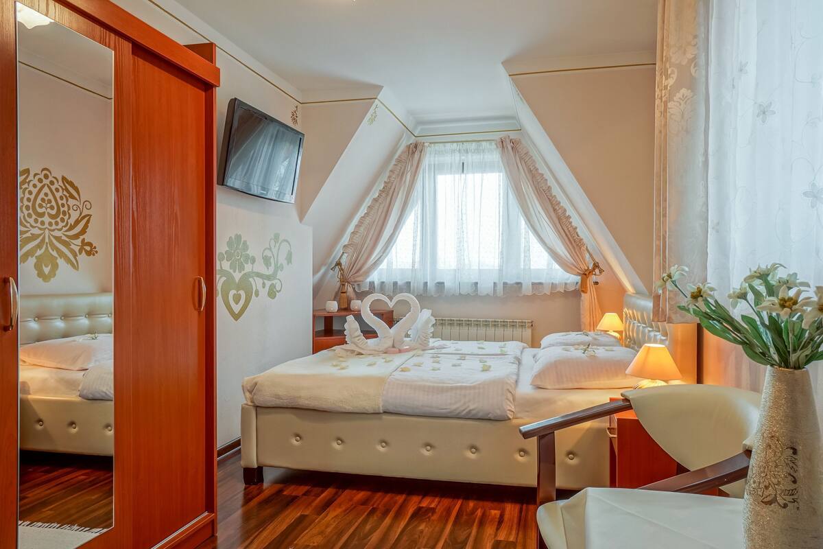 A bedroom with a bed, a TV, a window with curtains, a vase with flowers, and a wooden floor.