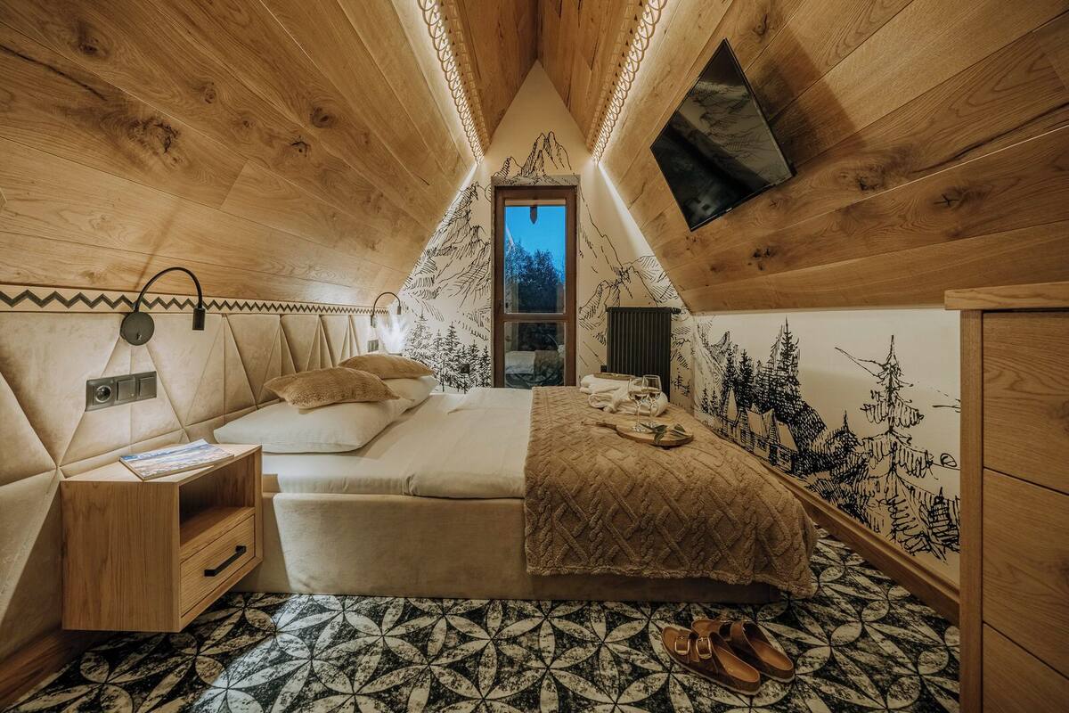 A cozy bedroom with a sloped ceiling, a bed with a decorative headboard, a nightstand, a dresser, and a window with a view of the outdoors.