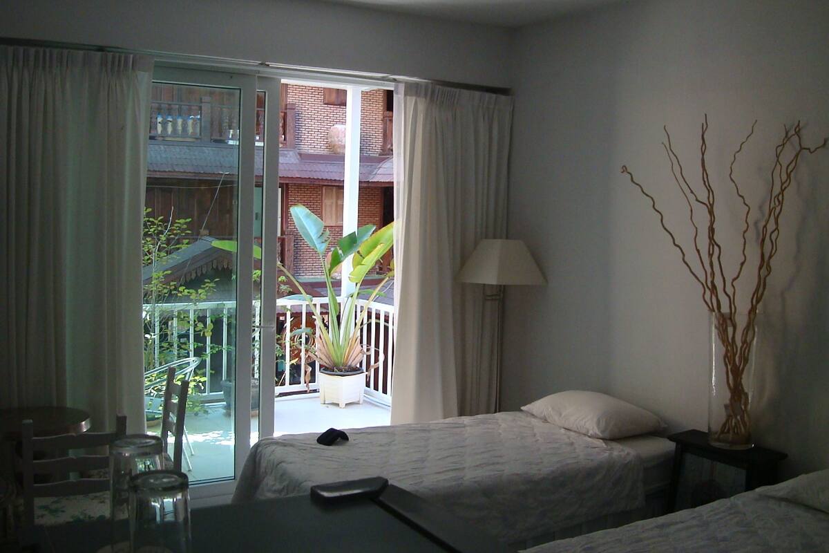 Habitación de hotel con una cama, una mesa y una silla. Cuenta con un balcón y vistas a un edificio y plantas.