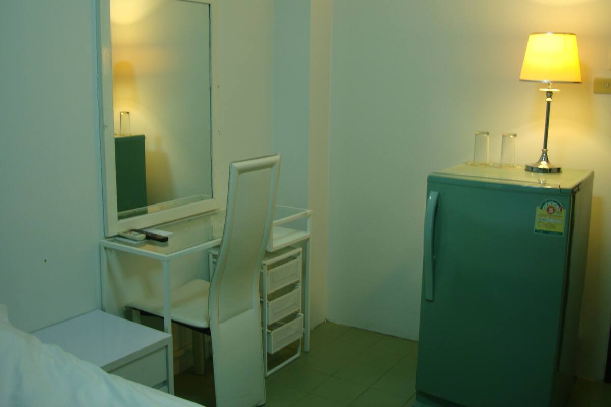 Habitación de hotel compacta con cama, escritorio con silla, espejo, refrigerador pequeño y lámpara de noche.