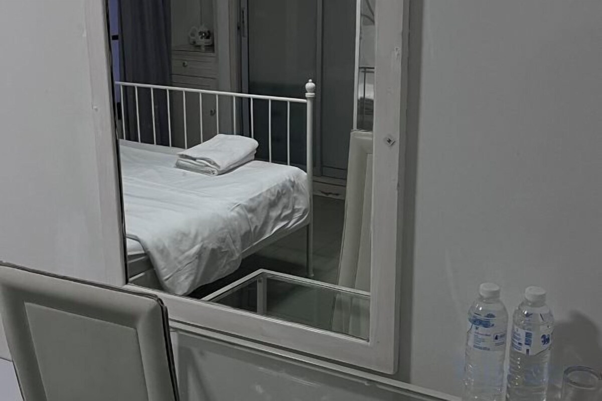 Una habitació d'hospital amb un llit individual, una taula de nit i un mirall que reflecteix l'habitació. Hi ha dues ampolles sobre la taula.