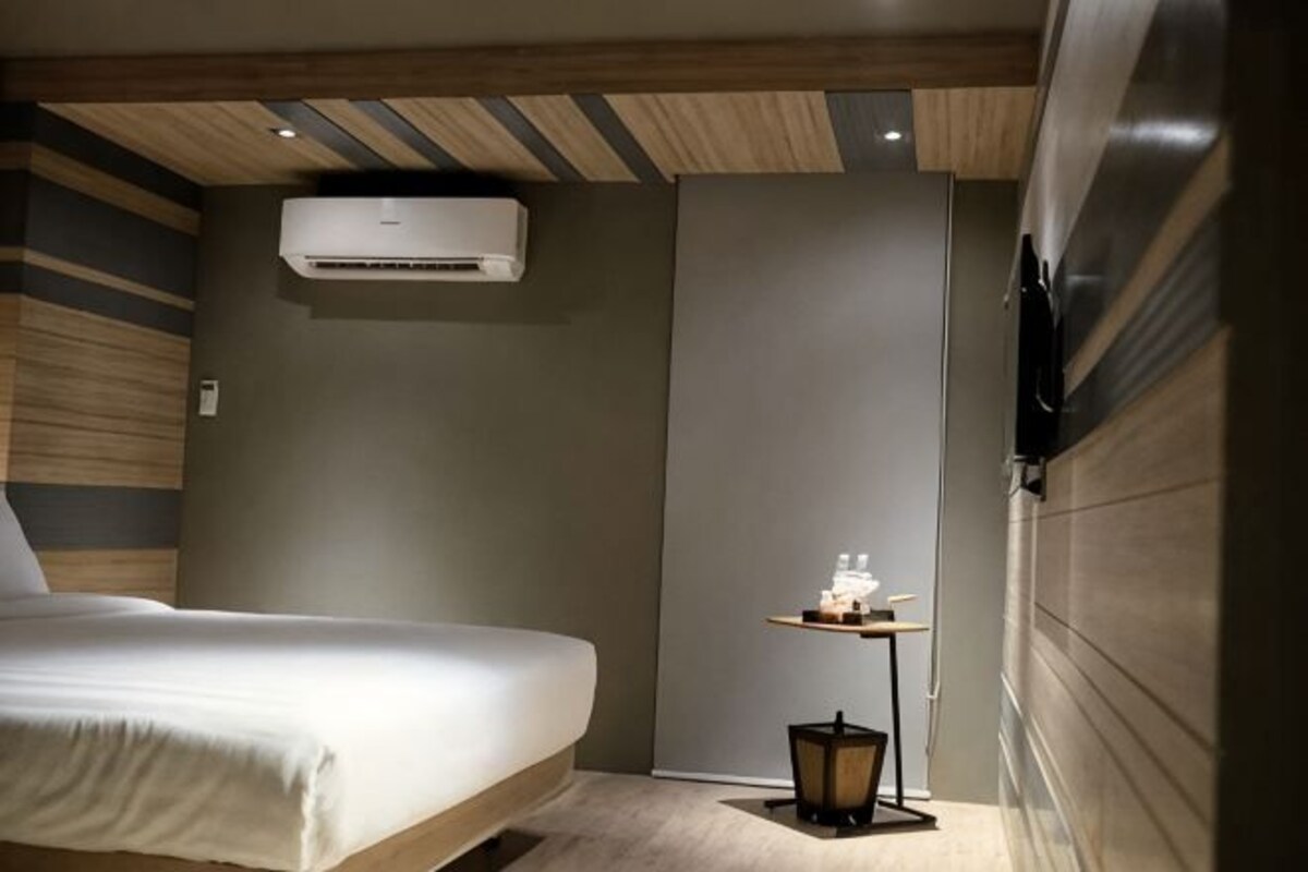 A hotel room with a bed, a small table with toiletries, an air conditioning unit, and a wall-mounted TV.