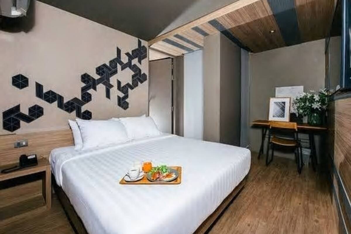 A modern hotel room with a neatly made bed, a wooden nightstand, a desk with a chair, and a geometric wall decoration.