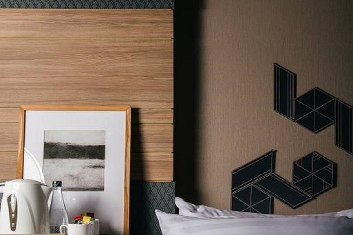 A hotel room with a bed, a nightstand, a framed picture, a kettle, and a mug.