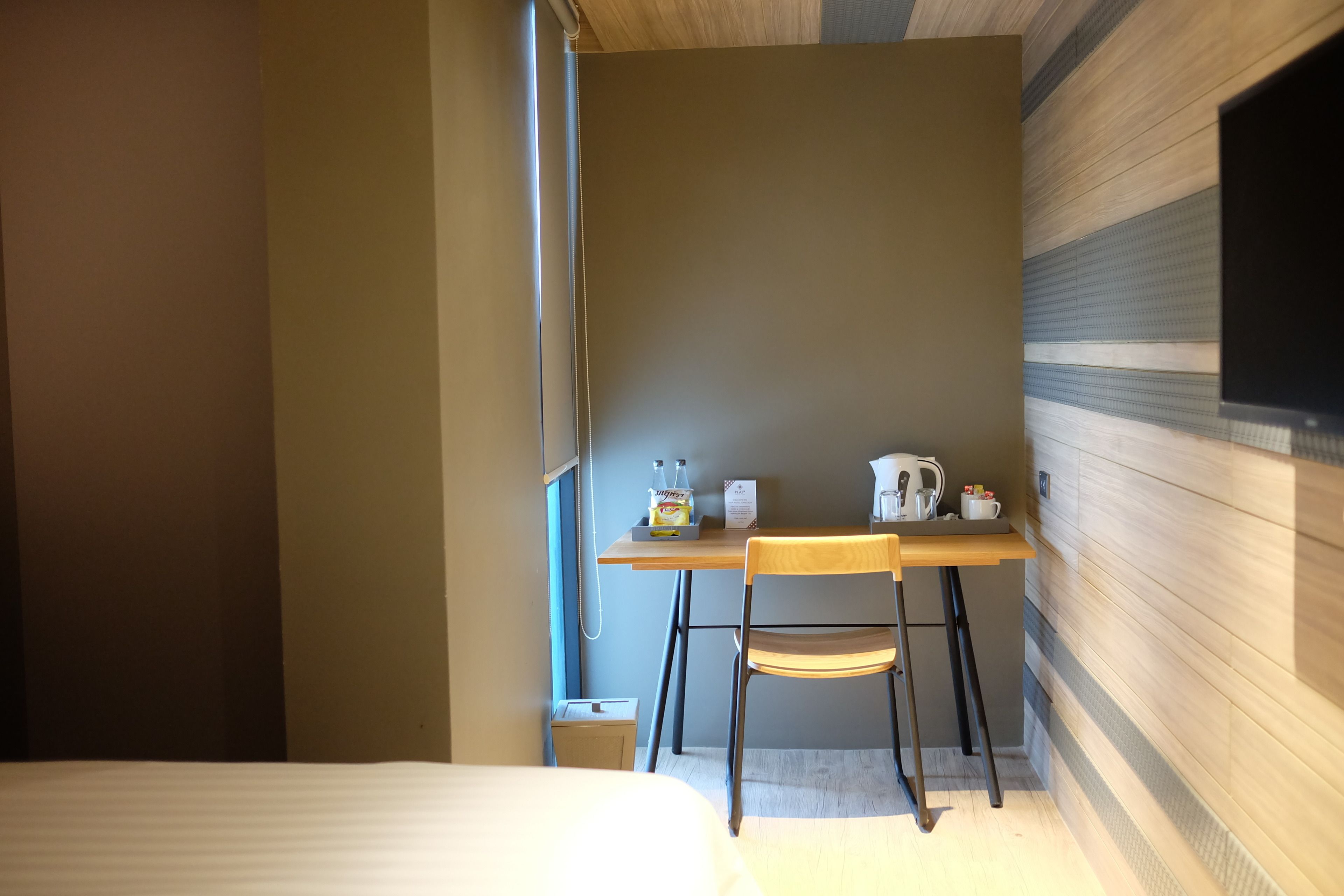 Een hotelkamer met een bed, een houten bureau met een stoel, een klein tafeltje met een waterkoker en een aan de muur bevestigde televisie.