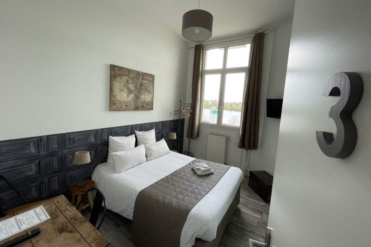 Ett hotellrum med en säng, ett skrivbord med en lampa, en karta på väggen och ett fönster med gardiner.