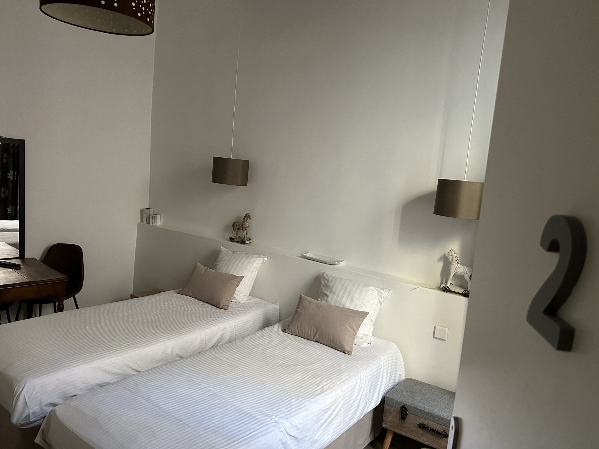A bedroom with two beds, a desk, and a chair. There are two wall-mounted lamps and a small shelf with decorative items.