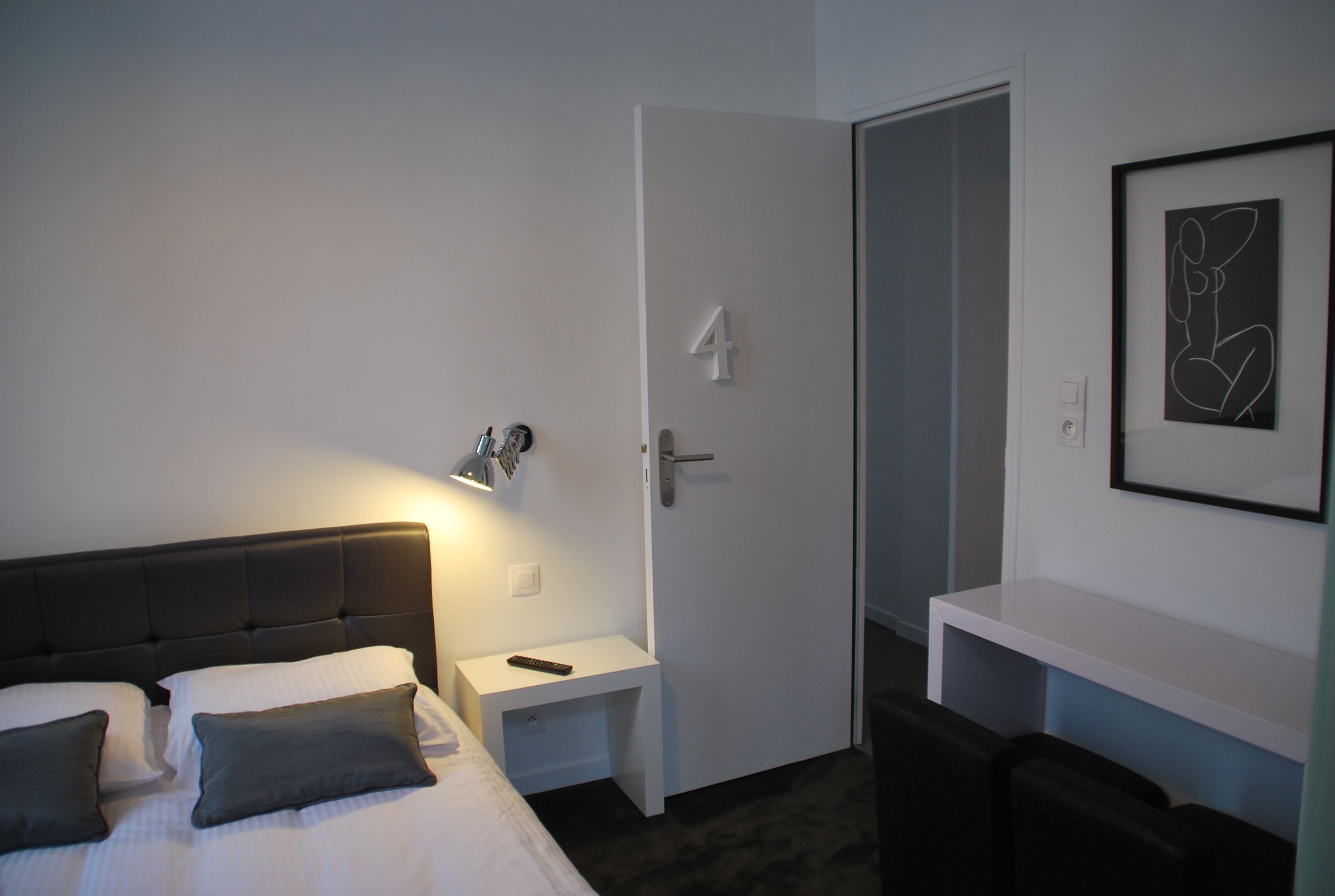 A hotel room with a bed, bedside table, a remote control, a door with a number, and a framed abstract art piece on the wall.