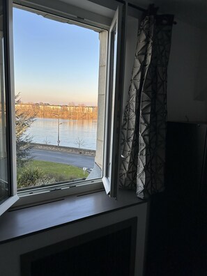 A window with a view of a body of water, a road, and buildings in the distance. - Rive Gauche (Saumur)