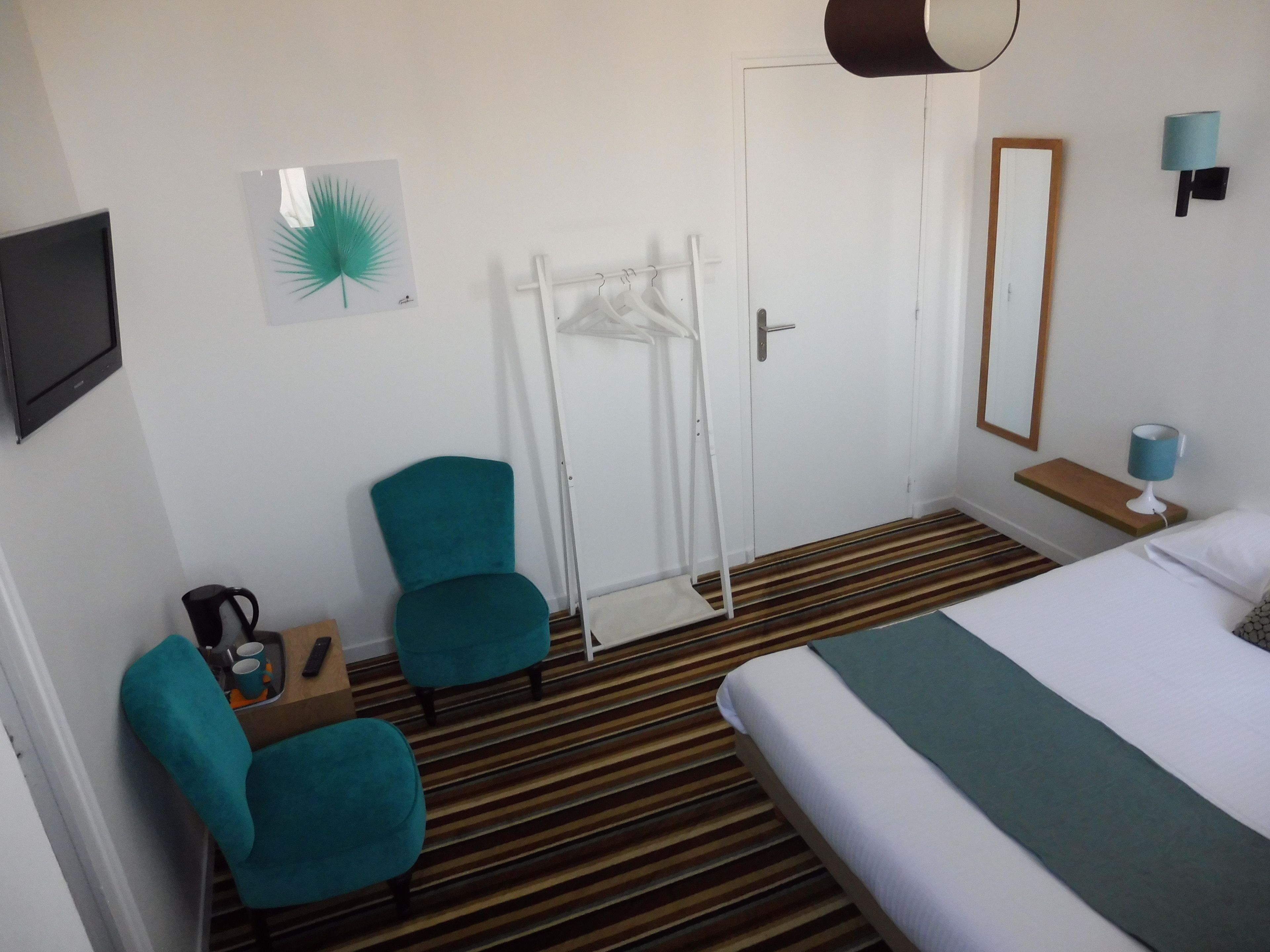 A hotel room with a bed, two teal chairs, a desk with a coffee maker, a TV, a door, a mirror, and a wall-mounted lamp.