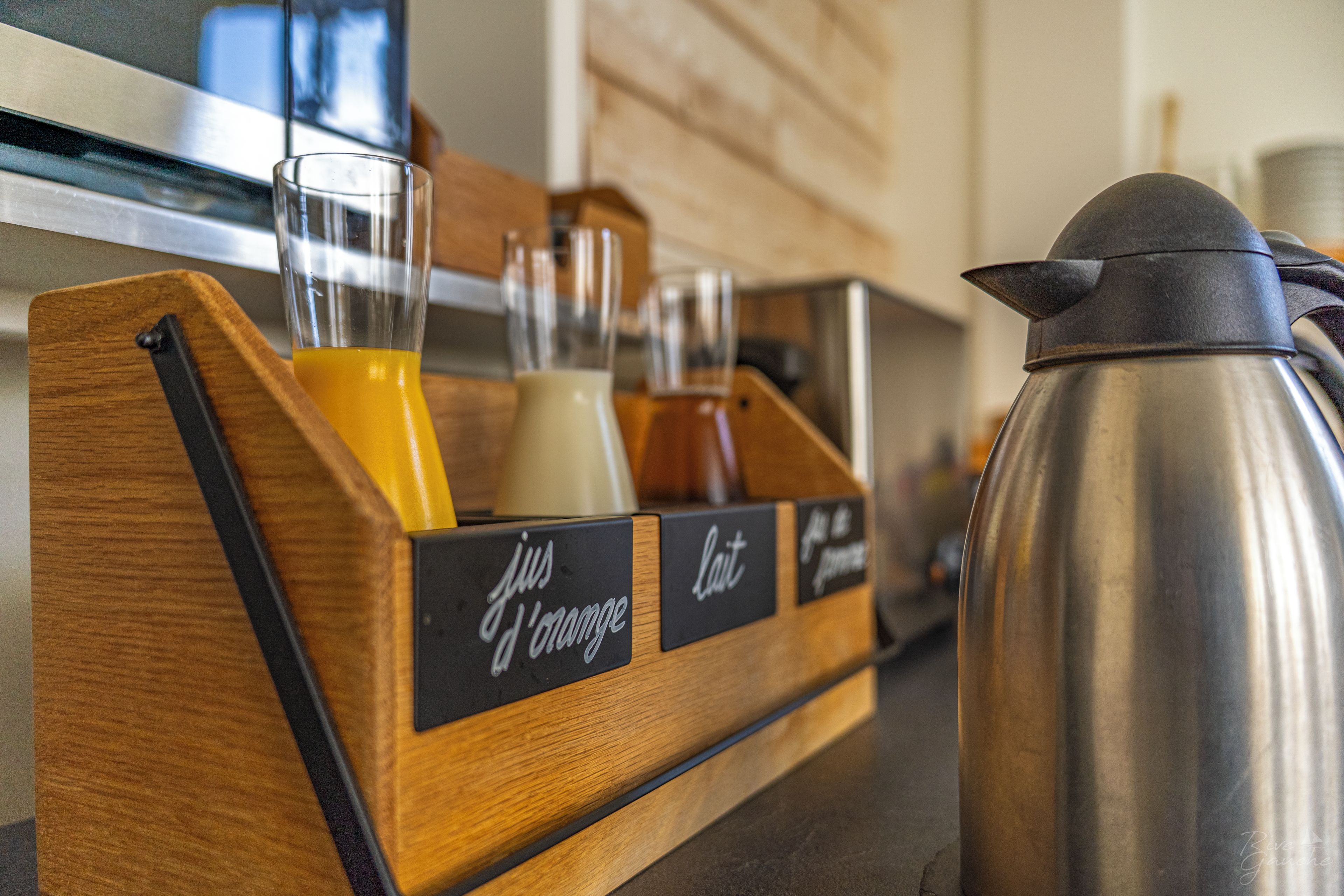 Een houten drankdispenser met glazen met etiketten voor sap en melk, en een roestvrijstalen koffiepot.