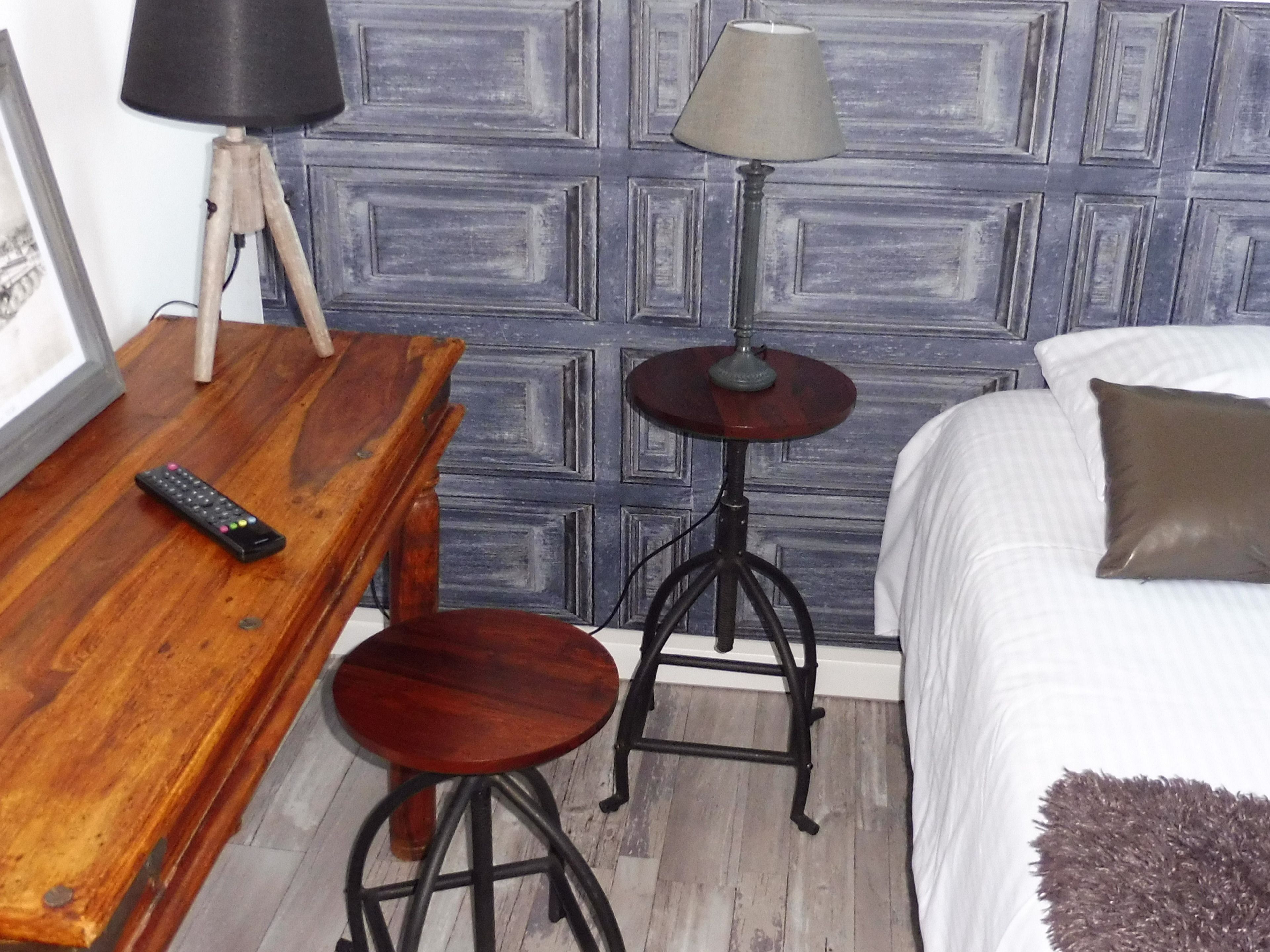 Une pièce comprenant un bureau en bois, une lampe, un tabouret, un lit avec un oreiller et un mur orné d’un motif géométrique.