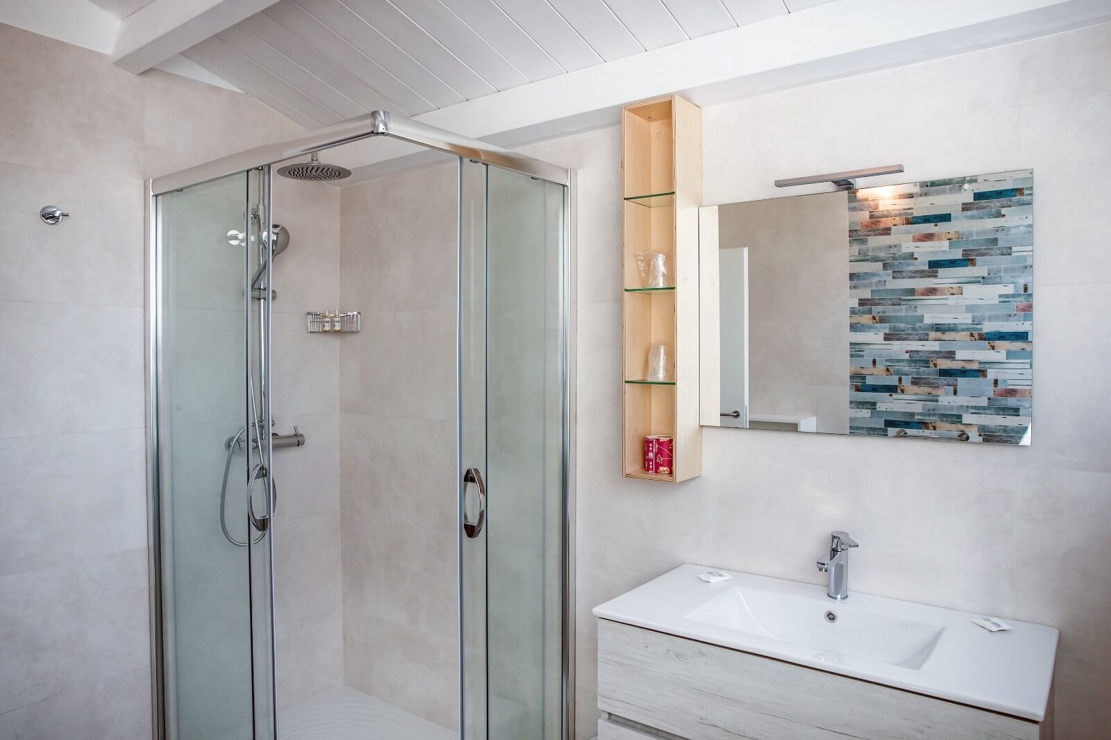 Een moderne badkamer met een douchecabine van glas, een houten plankje met toiletartikelen, een grote spiegel met een mozaïekachtergrond en een wit wastafelmeubel.